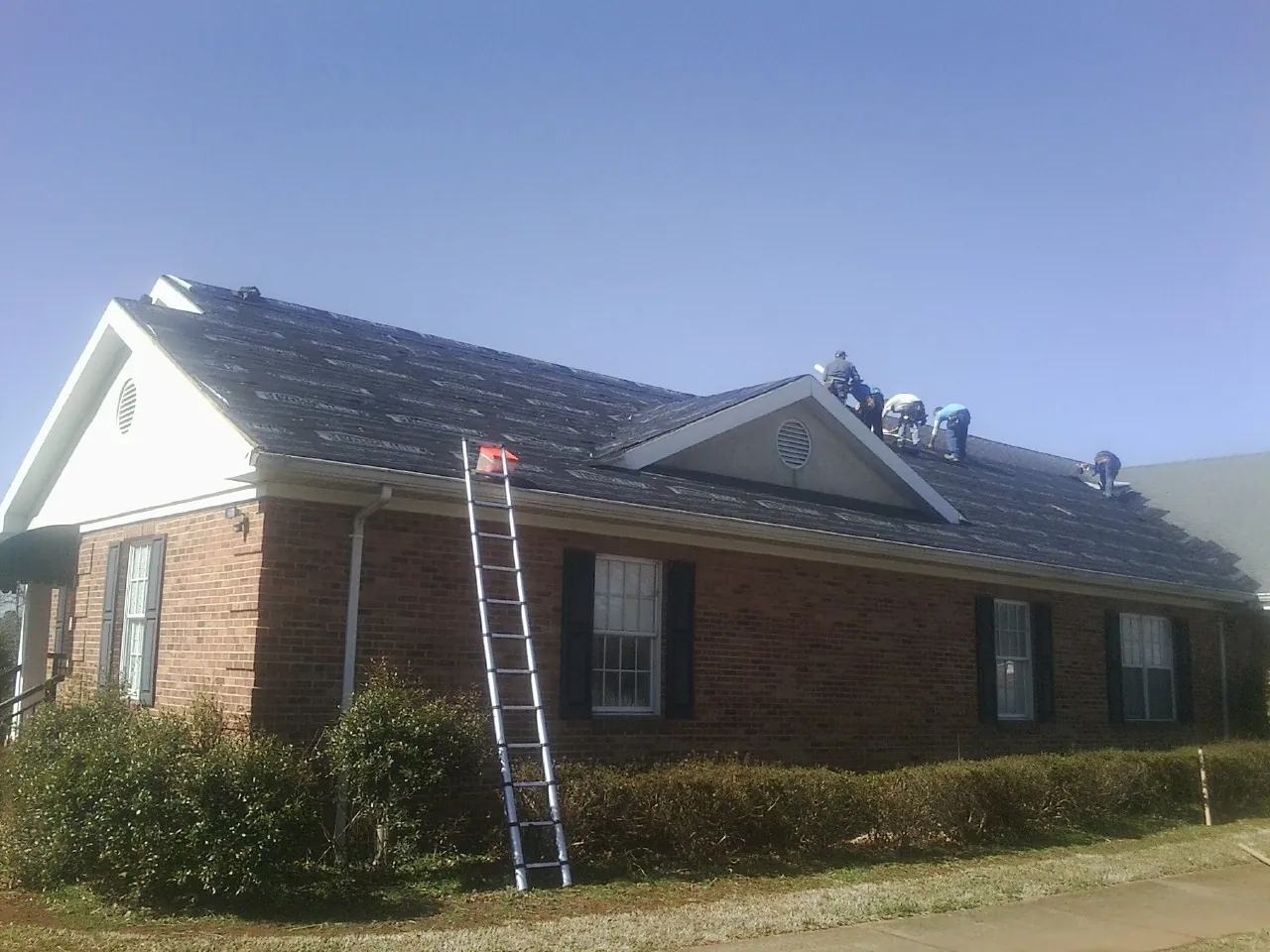 Crew tearing shingles off a brick ranch roof with a ladder set — Metal Roof Repair work by our crew in Ravenswood