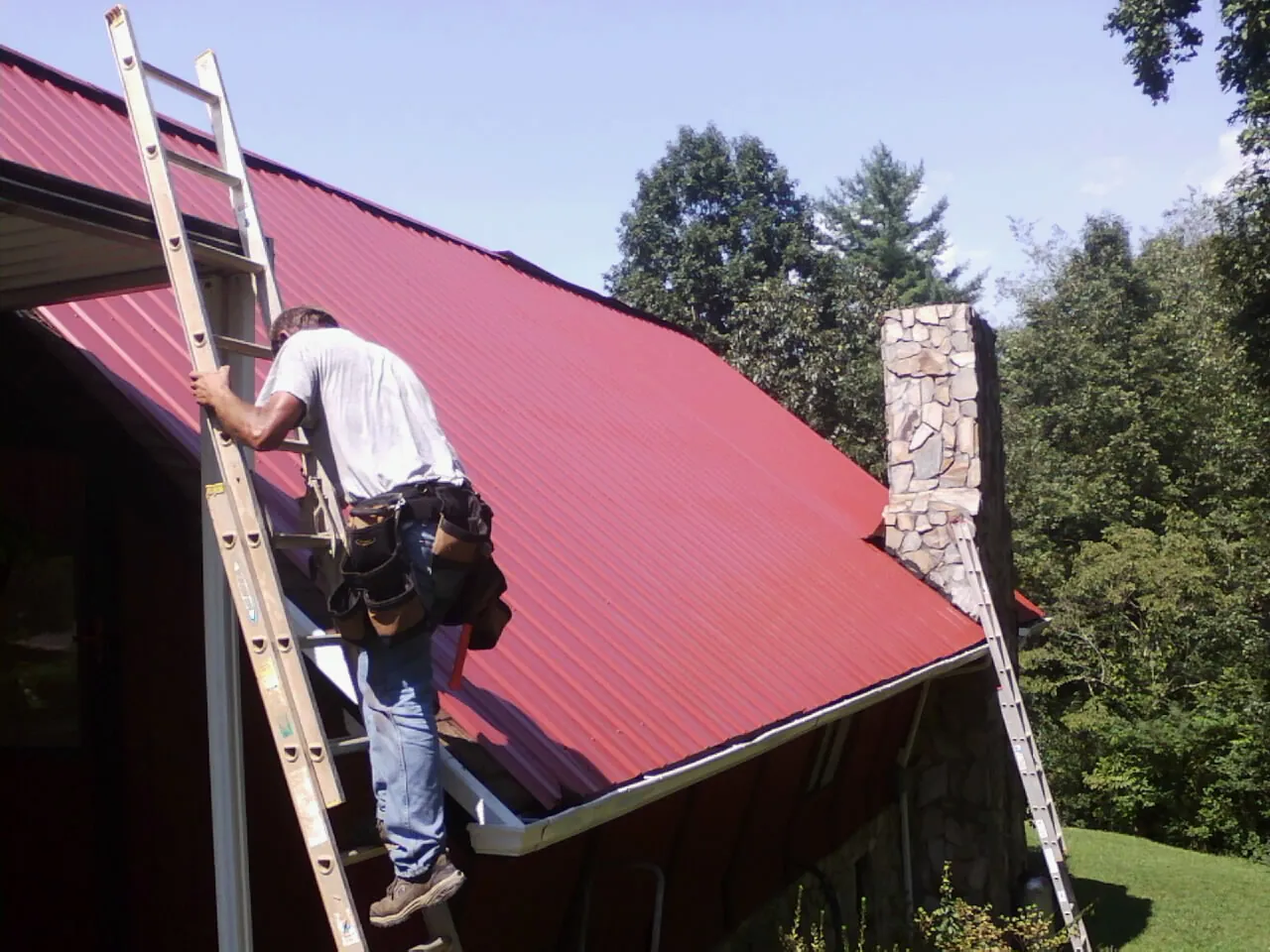 Roofer on a ladder beside a red metal roof and stone chimney — Soffit & Fascia Repair by Roofing Brothers in Ravenswood